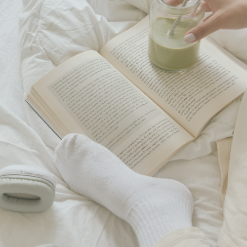 Woman sitting on a white puff blanket, socks resting on an open book, holding a green drink in a mason jar with a metal straw. Part of her tan pants and a small portion of her hand with a white-painted thumbnail are visible.