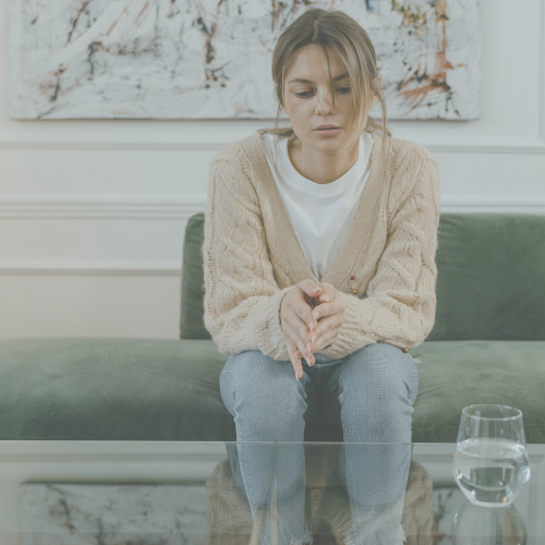 Woman sitting on a green couch, looking overwhelmed with messy hair in a bun, a glass cup of water on the coffee table in front of her.